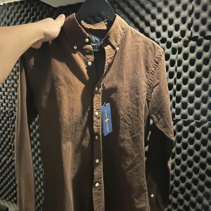 Brun skjorta Ralph Lauren - Snygg brun skjorta från Ralph Lauren i mjuk manchester med slim fit-passning. Säljer då jag fick en i present men har aldrig kommit till användning. Klassisk button-down krage, vita knappar och broderad logga på bröstet. Perfekt för dig som gillar stilrena och tidlösa plagg med lite edge.