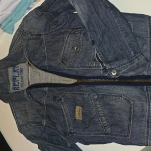 Blå jeansjacka från Replay - Säljer en snygg blå jeansjacka från Replay med tryck på ryggen och coola detaljer som dragkedja och knappar. Jackan har flera fickor framtill och klassisk krage. Perfekt för dig som gillar street och vill ha en jacka med attityd.