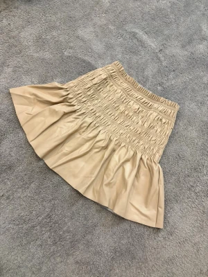 Beige kort kjol med volang - Supersnygg beige kort kjol med volangkant och smockad midja. Kjolen har en glansig yta som ger en cool look och är tillverkad i syntetmaterial som liknar skinn. Perfekt för dig som vill ha en trendig och bekväm kjol med extra volym.