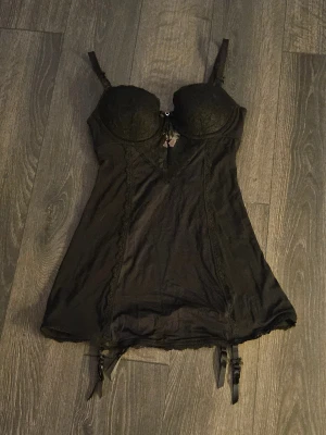 Svart spetsklänning från Hunkemöller - Supersnygg svart kortklänning från Hunkemöller med spetsdetaljer och justerbara axelband. Klänningen har vadderade kupor, dekorativ rosett framtill och fästen för strumpeband. Materialet är mjukt och följsamt med en lyxig känsla.