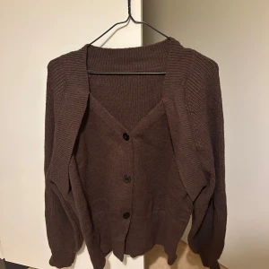 Brun stickad kofta med knappar - Mysig brun stickad kofta med v-ringning och tre svarta knappar framtill. Koftan har långa ärmar och ribbade detaljer vid krage, ärmslut och nederkant. Perfekt för lager-på-lager och chill dagar.
