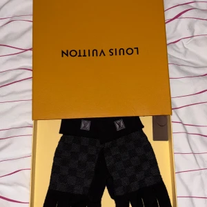 Svarta Louis Vuitton rutiga stickade vantar - Exklusiva svarta stickade handskar från Louis Vuitton med klassiskt rutig mönster och LV-logga på mudden. Snygg design med diskreta mörkgrå rutor och ribbade muddar. Perfekta för dig som vill ha både stil och värme under kalla dagar.
