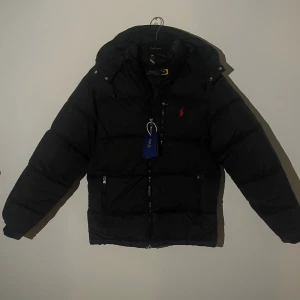 Ralph Lauren Jacka - Nu säljer jag en svart pufferjacka från Polo Ralph Lauren med huva och röd logga på bröstet. Jackan har två sidofickor med dragkedja och en kraftig frontzip. Materialet är syntet och dun, perfekt för kalla dagar. Klassisk och stilren look med quiltad design. Jackan är i nyskick och redo för en användare!!🙌❄️