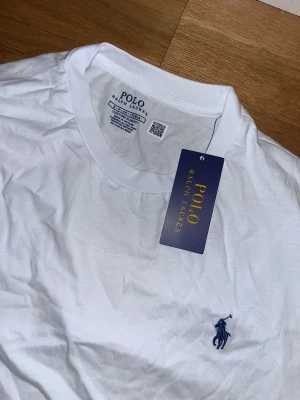 Vit t-shirt från Polo Ralph Lauren - Vit t-shirt från Polo Ralph Lauren med klassisk rund hals och korta ärmar. Slim fit-modell i mjuk bomull med den ikoniska blå broderade loggan på bröstet. Perfekt basic med stilren design och skön passform.