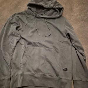 Grå hoodie från Jack & Jones - Säljer en enkel och snygg grå hoodie från Jack & Jones. Tröjan har huva med snörning, stor magficka och ribbade muddar vid ärmslut och nederkant. 
