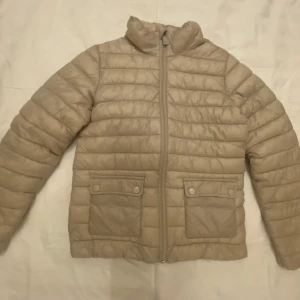 Beige pufferjacka med fickor - Snygg beige pufferjacka med horisontella quiltade sömmar och två stora fickor framtill med lock och knappdetaljer. Jackan har dragkedja hela vägen upp och en hög krage som håller dig varm. Ser ut att ha en tråkig färg men endast i kamera. Har en liten fläck som knappt syns på armen baksidan. 