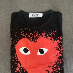 Svart t-shirt Comme des Garçons Play - Svart t-shirt från Comme des Garçons Play med stort rött hjärta och ögon framtill, designad med coolt färgstänksmönster. Rund hals och klassisk passform, perfekt för dig som gillar statement-plagg och streetstyle.