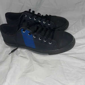 Saint Gabriel Sneakers - Snygga svarta Saint Gabriel sneakers med en blå diagonal detalj på sidan. Skorna har klassisk snörning, rund tå och platt sula med vit rand längs kanten. Tillverkade i textilmaterial och passar perfekt till en avslappnad streetstyle-look.