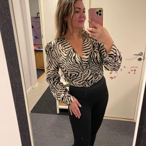 Zebramönstrad body-blus med v-ringning - Snygg body-blus med zebramönster i svart och beige, tillverkad i ett glansigt material som ger en lyxig känsla. Blusen har v-ringad hals, långa ärmar och en loose passform. Perfekt för dig som vill sticka ut med ett djuriskt print.helt ny