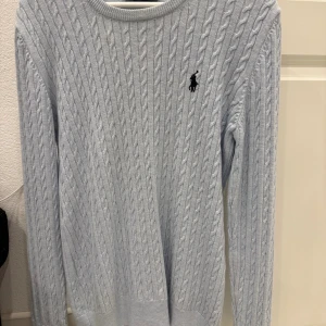 Ljusblå stickad tröja från Ralph Lauren - Snygg ljusblå stickad tröja från Ralph Lauren med klassiskt kabelstickat mönster och broderad logga på bröstet. Rund halsringning och långa ärmar gör den perfekt för kyliga dagar. Passar dig som gillar stilren och tidlös design.