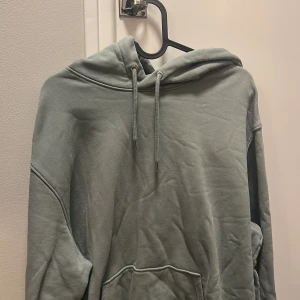 Grå hoodie med snörning - Snygg grå hoodie med huva och snörning framtill. Tröjan har en klassisk känguruficka och långa ärmar, perfekt för chill dagar. Mjuk bomullskänsla och relaxed passform gör den superbekväm att bära.