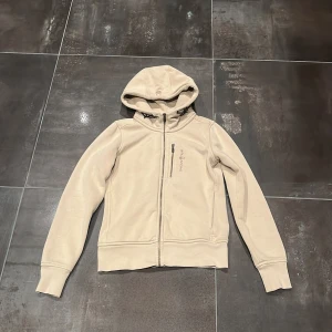 Beige hoodi från Sail Racing  - Snygg beige hoodi från Sail Racing i storlek XS. Jackan har huva med snörning, hel dragkedja framtill och en bröstficka med zip. Mjuk insida i fleece och stilren design, perfekt för kyliga dagar. Långärmad och med ribbade muddar för extra komfort.