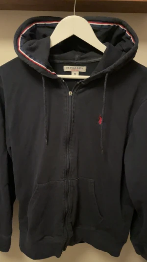 Mörkblå hoodie - Snygg mörkblå hoodie från U.S. Polo Assn. med dragkedja och huva med röd, vit och blå kant. Tröjans skick är 8/10 förutom att dragkedjan är trasig se bild 2. Står M men passar S. Snabb affär kan priset sänkas.