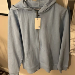Ljusblå sip hoodie från lager 157 - Snygg ljusblå hoodie från 157 med hel dragkedja framtill och två stora fickor. Tröjan har huva med snörning och ribbade muddar vid ärmslut och nederkant. Perfekt för chill dagar och enkel att matcha med jeans eller mjukis.