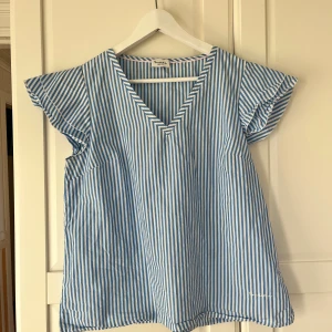 Blårandig blus från Bondelid - Säljer en fräsch blus från Bondelid med blå och vita ränder. Blusen har v-ringad hals, korta volangärmar och är tillverkad i mjuk bomull. Perfekt för dig som gillar en luftig och chill stil med lite extra detaljer på ärmarna.