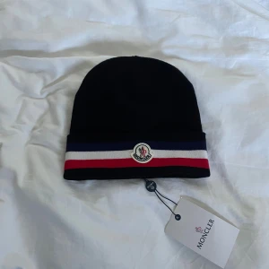Svart M mössa med ränder - Svart mössa från M med klassiska ränder i blått, vitt och rött på uppviket. Märkets logga sitter framtill och materialet är mjuk stickad bomull. Perfekt för dig som vill ha en clean och stilren look med lite lyxig vibe.