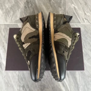 Valentino Rockrunner sneakers camouflage - Snygga Valentino Rockrunner sneakers med grön, beige och svart camouflage-mönster. Skorna har detaljer i mocka och mesh, svarta hälpartier och klassiska nitar på sidan av sulan. Platt sula och rund tå, med snörning framtill. Perfekt för dig som gillar streetstyle.