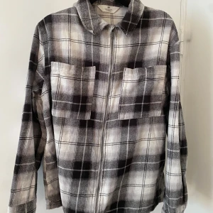 Rutig overshirt jacka från H&M - Snygg overshirt jacka från H&M i svart, grå och vit rutig design. Jackan har två stora bröstfickor, dragkedja framtill och klassisk krage. Perfekt för lager-på-lager och chill stil. Materialet är mjukt och känns som bomull. Passar dig som gillar street och casual.