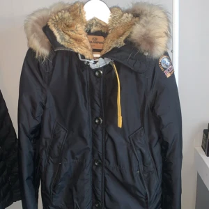 Svart Parajumpers parkajacka med päls - Svart parkajacka från Parajumpers med fluffig päls runt huvan och insidan. Jackan har gul detalj vid halsen, flera fickor och knappar framtill. Märkespatch på ärmen och grov dragkedja. Perfekt för kalla dagar och riktigt snygg siluett.