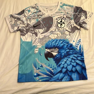 Nike Brasil CBF blå papegojtröja - Unik fotbollströja från Nike med Brasil CBF-logga. Tröjan har ett färgstarkt tryck med en blå papegoja, blommor och grafiska detaljer i vitt, grått och blått. Kortärmad modell i lätt, ventilerande material. Perfekt för dig som gillar djärva mönster och sportig stil.