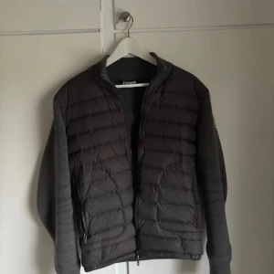 Moncler cardigan - Säljer en grå pufferjacka från Moncler med quiltad framsida och mjuka stickade ärmar. Jackan har två fickor med dragkedja och Moncler-logga på ärmen. Perfekt för dig som vill ha en snygg och bekväm jacka med streetkänsla. Storlek L men sitter som M