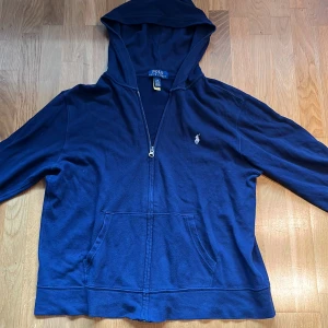 Marinblå hoodie från Polo Ralph Lauren - Snygg marinblå hoodie från Polo Ralph Lauren med dragkedja och klassisk broderad logga på bröstet. Tröjan har huva, känguruficka och är tillverkad i mjuk bomull. Perfekt för en chill och stilren look. Strlk L (14-16)
