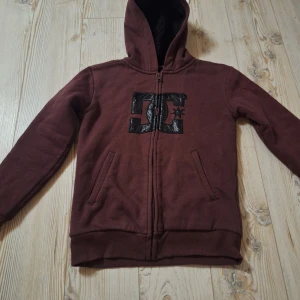 Röd hoodie från DC med huva - ⚠️OBS! Storleken är BARN S!⚠️            Säljer en vinröd hoodie från DC med stor svart logga på bröstet. Tröjan har dragkedja, två fickor fram och mysigt svart fleecefoder på insidan. Perfekt för kyliga dagar och har en skön, normal passform. Snygg och enkel att matcha med jeans eller joggers.