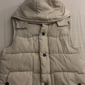 Beige dunväst med huva - Snygg beige dunväst med stor huva och svarta knappar framtill, man kan även ta av huvan om man vill det. Perfekt att slänga över en hoodie när det är kyligt ute. Hör av er vid intresse 