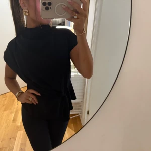 Svart omlottopp med knyt i midjan - Stilren svart topp med hög hals och korta ärmar. Toppen har en snygg omlottdesign och knyt i midjan som ger en modern siluett. Perfekt att styla med smycken och accessoarer för en trendig look.