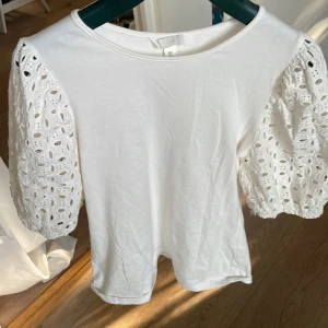 Vit blus med broderade puffärmar H&M - Superfin vit blus från H&M med broderade puffärmar i spets och rund halsringning. Ärmarna är korta och har ett snyggt hålmönster som ger en romantisk vibe. Perfekt att styla med jeans eller kjol för en fräsch look.