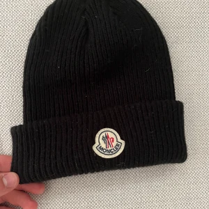 Svart  mössa i ull - Svart ribbstickad mössa  med klassisk uppvikt kant och broderad logotyp framtill. Snygg och enkel design som passar till allt, tillverkad i mjuk ull som håller dig varm under kalla dagar.