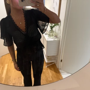 Svart prickig blus med spetskant - Säljer en svart blus i transparent material med prickigt mönster. Blusen har v-ringning med fin spetskant, korta vida ärmar och knyt i midjan. Perfekt för dig som gillar feminina detaljer och en luftig känsla.