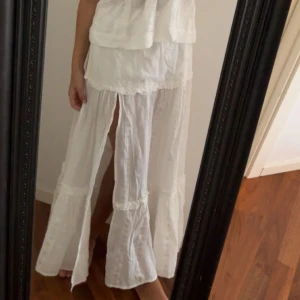 Vit långkjol med slits och volang - Superfin vit långkjol med volangdetaljer och en snygg slits framtill. Kjolen har en luftig och flowy känsla, perfekt för varma dagar. Passar dig som gillar en romantisk och bohemisk stil.