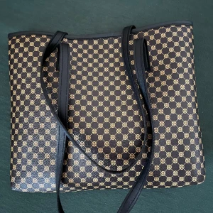 Svart och guldmönstrad axelväska - Snygg axelväska i svart med guldigt geometriskt mönster. Väskan har långa svarta axelband och är rymlig med öppen topp. Perfekt för dig som vill ha en stilren och trendig väska med plats för allt du behöver.