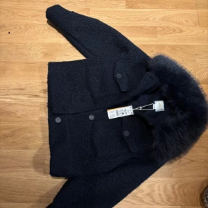 Svart jacka med pälsdetalj från Ginatricot - Snygg mörkblå jacka från Ginatricot med en fluffig pälsdetalj runt kragen. Jackan har knappar framtill och två stora fickor. Perfekt för kyligare dagar med sin stilrena design. Använd några gånger 
