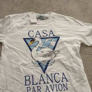 Vit t-shirt Casablanca med svanprint - Casablanca t-shirt i vitt med stor grafisk svan och blå text 'Casa Blanca Par Avion' på framsidan. Klassisk rund hals och korta ärmar. Materialet är mjuk bomull och passformen är normal. Perfekt för dig som gillar statement-plagg och coola prints.