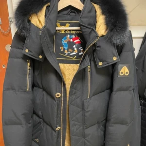 Svart dunjacka Moose Knuckles - Snygg svart dunjacka från Moose Knuckles med gulddetaljer, pälskantad huva och flera dragkedjefickor. Jackan har quiltad design, guldig insida och logga på ärmen. Perfekt för kalla dagar när du vill hålla stilen.