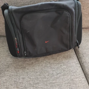 Svart datorväska från Nike - Säljer en svart datorväska från Nike med röd logga framtill. Väskan har justerbar axelrem, flera dragkedjefack och vadderat innerfack för laptop. Perfekt för skolan eller jobbet när du vill ha med dig datorn på ett smidigt sätt.