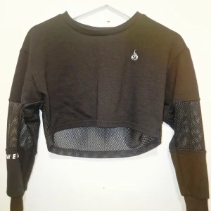 Svart croppad meshtröja från Ryderwear  - Svart croppad långärmad tröja med meshdetaljer på rygg, ärmar och nederkant. Perfekt för träning, dans eller en sportig look. Använd max 3 gånger.