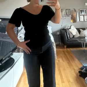 Svart ribbad topp med knyt i ryggen - Svart topp med kort ärm och v-ringning framtill. Toppen är ribbad och har snygg knyt-detalj i ryggen som ger en extra touch. Figurnära passform och mjukt material, perfekt till jeans eller kjol.