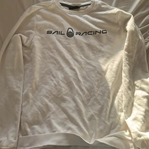 Vit Sail Racing sweatshirt - Vit långärmad sweatshirt från Sail Racing med svart logga framtill och liten logga i nacken. Klassisk rund halsringning och mjukt material som känns skönt mot huden. Perfekt för dig som gillar stilren och sportig look.