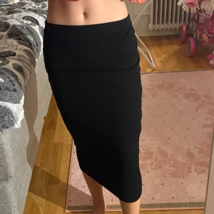 Svart långkjol i stretchigt tyg - Enkel och stilren svart långkjol med tight passform och mjukt, stretchigt material. Kjolen har en rak siluett och slutar strax under knäet. Perfekt för dig som gillar minimalistisk och clean stil. Den är ribbad