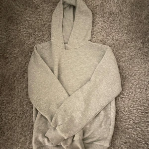 Grå hoodie från Gina Tricot med tryck - Grå hoodie från Gina Tricot med rosa texten 'LUXUEUSE BONHEUR' och en liten rosett på ryggen. Klassisk modell med huva och ribbade muddar. Perfekt för dig som gillar enkel stil med en söt detalj. Mjuk och skön i bomullsmix.