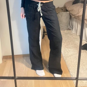 Svarta linnebyxor från Roxy med snörning - Svarta linnebyxor från Roxy med avslappnad passform och breda ben. Byxorna har resår i midjan och vit snörning framtill, samt fickor både fram och bak. Perfekta för chill dagar eller när du vill ha en skön och luftig stil.