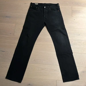 Svarta Levi's 501 jeans W32 L34 - Klassiska svarta Levi's 501 jeans med raka ben och femficksdesign. Jeansen har knappgylf, silverfärgade och kopparfärgade detaljer samt den ikoniska läderpatchen bak i midjan. Perfekta för dig som gillar en tidlös och clean look.