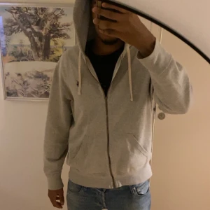 Grå Polo Ralph Lauren hoodie med dragkedja - Säljer en stilren grå hoodie med dragkedja och huva. Tröjan har vita snören, fickor framtill och en klassisk passform. Perfekt för chill dagar eller när du vill ha en enkel och snygg look.