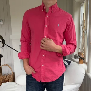 Rosa skjorta från Polo Ralph Lauren - Snygg rosa skjorta från Polo Ralph Lauren i slim fit-modell. Skjortan har klassisk button down-krage, långa ärmar och knappar framtill. Broderad logga i ljusblått på bröstet. Perfekt för dig som gillar stilrena och färgstarka plagg.