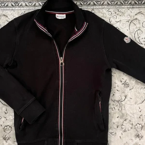 Svart Moncler kofta med dragkedja - Svart kofta från Moncler med hel dragkedja och ribbade muddar. Kragen har snygga ränder i vitt och rött, och Moncler-loggan sitter på ärmen. Perfekt för dig som gillar stilrena och exklusiva plagg.