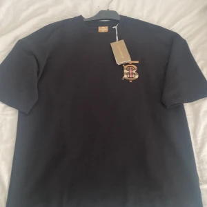 Svart Burberry t-shirt med tryck - Svart t-shirt från Burberry i bomull, med klassiskt rund hals och korta ärmar. På bröstet finns ett broderat Burberry-monogram med rutigt mönster och röd detalj. På ryggen finns ett gult texttryck med motiverande citat. Perfekt för dig som gillar stilrena designer med en twist. Pris kan diskuteras!!