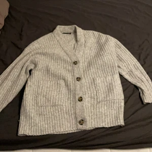 Ljusgrå stickad kofta från 157 - Mysig ljusgrå stickad kofta från 157 i storlek M. Koftan har grov ribbstickning, stora framfickor och snygga knappar framtill. Perfekt att slänga på sig när det är kyligt ute. Passar dig som gillar chill och enkel stil.